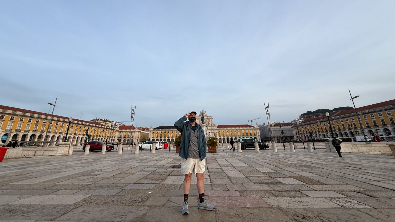 A photo of Dave in a wide plaza in Lisbon. There are yellow buildings and an arch in the background.